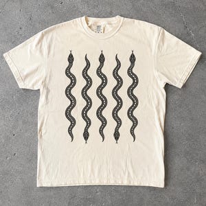 May include: A cream-colored short-sleeve t-shirt featuring a graphic print of five vertical, wavy black snakes arranged in a parallel pattern across the center of the chest. The snakes have detailed, repeating geometric scale patterns along their bodies.
