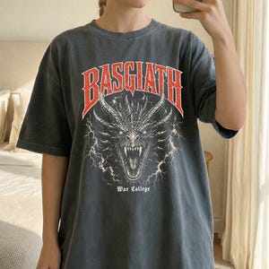 Puede incluir: Camiseta gris oscuro con aspecto desgastado. La parte delantera presenta un gr&aacute;fico rojo y blanco de una cabeza de drag&oacute;n con la palabra "BASGIATH" arriba y "War College" abajo. La camiseta tiene mangas cortas y cuello redondo.