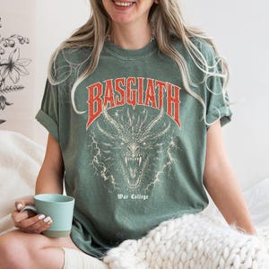 Puede incluir: Camiseta verde salvia con un dise&ntilde;o de drag&oacute;n y la palabra "BASGIATH" en rojo encima. El texto "War College" est&aacute; debajo del drag&oacute;n. La camiseta tiene las mangas dobladas. Una persona lleva la camiseta y sostiene una taza azul.