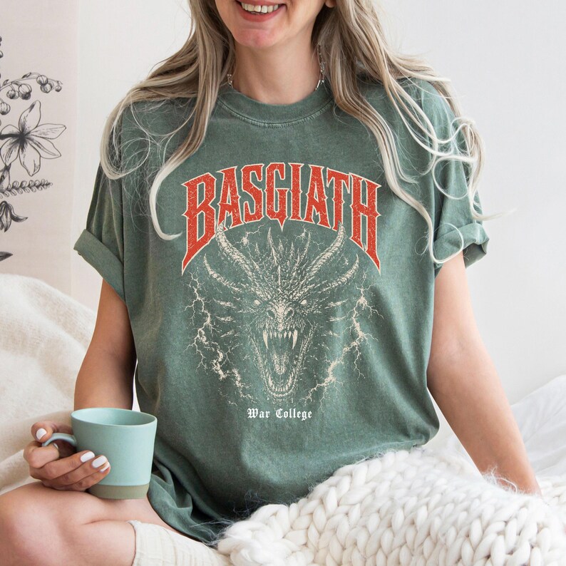 Puede incluir: Camiseta verde salvia con un dise&ntilde;o de drag&oacute;n y la palabra "BASGIATH" en rojo encima. El texto "War College" est&aacute; debajo del drag&oacute;n. La camiseta tiene las mangas dobladas. Una persona lleva la camiseta y sostiene una taza azul.