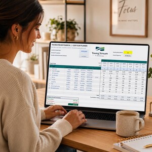 May include: A woman using a laptop displaying a cash flow planner. The screen shows a spreadsheet with financial data. A mug, notebook, and pen are on the wooden desk. The laptop screen displays the text "Young Stream Financial".