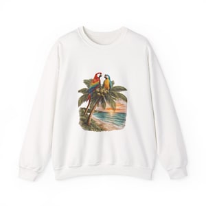 May include: White sweatshirt featuring a colorful graphic of two parrots perched on a palm tree with a beach scene in the background. The parrots have red, blue, yellow, and green feathers. The beach scene includes a sunset over the ocean.