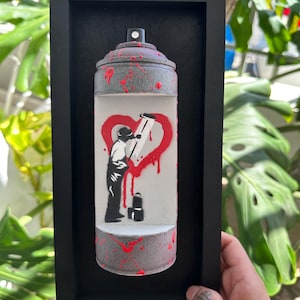 Puede incluir: Obra de arte enmarcada que presenta un diseño de bote de pintura en aerosol. El bote es gris con salpicaduras rojas y una boquilla negra. En el interior, una silueta negra pinta un corazón rojo. La obra está sobre un fondo negro dentro de un marco negro.