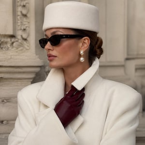 May include: A cream-colored pillbox hat, a cream-colored wool coat, dark burgundy leather gloves, black oval-frame sunglasses, and gold-toned drop earrings featuring a square setting with a dangling white pearl bead.