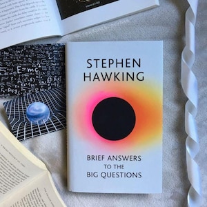 Op de afbeelding: Een boek van Stephen Hawking getiteld "Korte antwoorden op de grote vragen". De omslag heeft een zwarte cirkel met een roze en oranje gradiënt. Andere boeken en een wit lint zijn zichtbaar op de achtergrond.