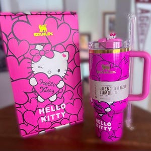 Puede incluir: Un vaso Stanley Quencher H2.0 rosa Hello Kitty con una caja a juego. El vaso presenta un asa, una pajita y un dise&ntilde;o de Hello Kitty con corazones. La caja y el vaso tienen el texto "Hello Kitty" y "Stanley".