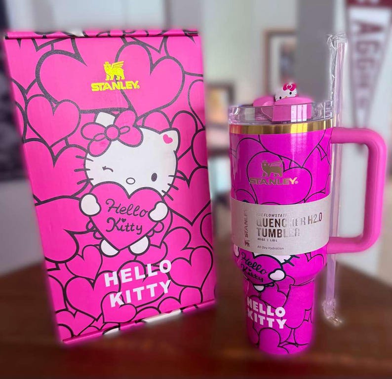 Puede incluir: Un vaso Stanley Quencher H2.0 rosa Hello Kitty con una caja a juego. El vaso presenta un asa, una pajita y un dise&ntilde;o de Hello Kitty con corazones. La caja y el vaso tienen el texto "Hello Kitty" y "Stanley".