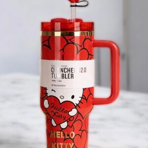Puede incluir: Vaso rojo de Hello Kitty con asa, pajita y adorno en forma de coraz&oacute;n. El vaso presenta un patr&oacute;n de corazones repetidos y un gr&aacute;fico de Hello Kitty sosteniendo un coraz&oacute;n. Tambi&eacute;n se ve el texto "QUENCHER 12.0 TUMBLER" y "HELLO KITTY".