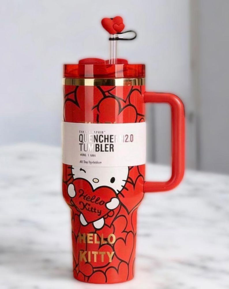 Puede incluir: Vaso rojo de Hello Kitty con asa, pajita y adorno en forma de coraz&oacute;n. El vaso presenta un patr&oacute;n de corazones repetidos y un gr&aacute;fico de Hello Kitty sosteniendo un coraz&oacute;n. Tambi&eacute;n se ve el texto "QUENCHER 12.0 TUMBLER" y "HELLO KITTY".