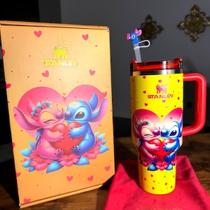 May include: A yellow Stanley tumbler with a red handle and lid, featuring a cartoon couple holding a heart. The tumbler is decorated with small red hearts and the Stanley logo. A matching box and red bag are also present.