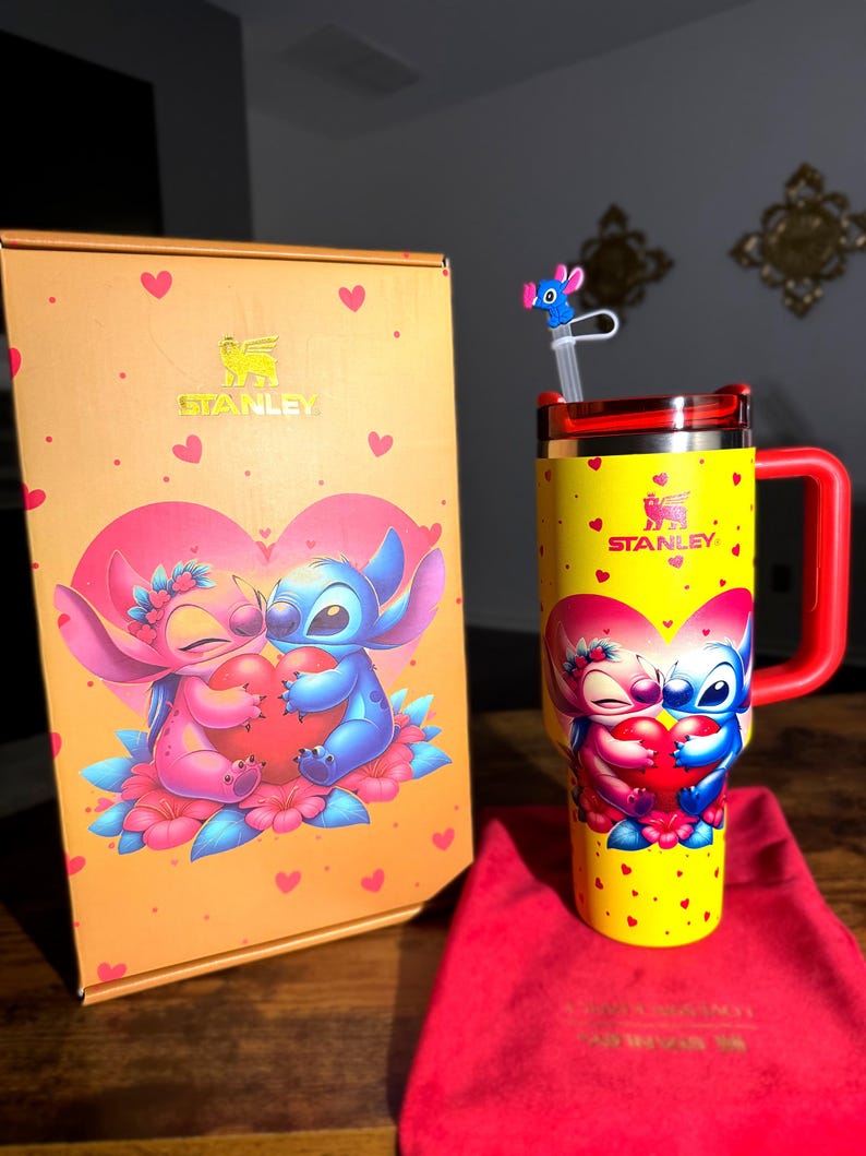 May include: A yellow Stanley tumbler with a red handle and lid, featuring a cartoon couple holding a heart. The tumbler is decorated with small red hearts and the Stanley logo. A matching box and red bag are also present.