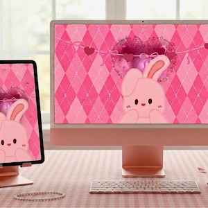 May include: A pink desktop computer and tablet display a matching wallpaper featuring a cartoon bunny and heart design. The setup includes a keyboard, mouse, and a decorative cup on a pink checkered surface.