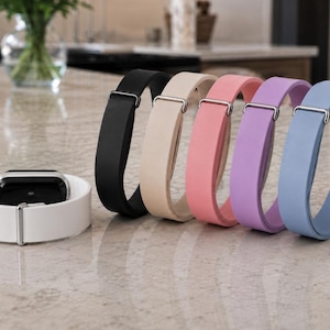May include: A collection of six silicone watch bands featuring a smooth, matte finish. One white band is attached to a black watch face, while five additional bands in black, beige, pink, lavender, and light blue stand upright with silver buckles.