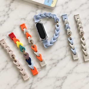 May include: A collection of replacement watch bands featuring a linked chain design with teardrop-shaped segments. The bands come in various colors including light blue, orange, yellow, green, purple, dark blue, and beige, with one light blue band attached to a black fitness tracker.