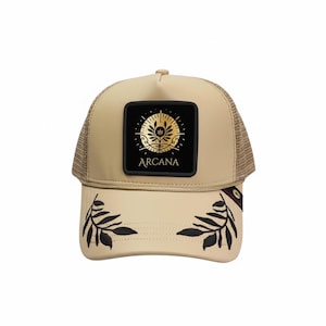 May include: Tan trucker hat with a mesh back and a curved brim. The front features a black square patch with gold floral design and the word "ARCANA". Black leaf embroidery adorns the brim.