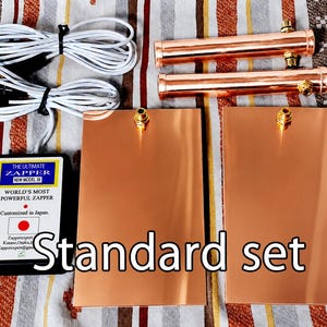 May include: A standard set including a black electronic device labeled The Ultimate Zapper New Model XE, two rectangular copper plates, two cylindrical copper tubes, and two white electrical cables with gold-colored connectors and black plugs.