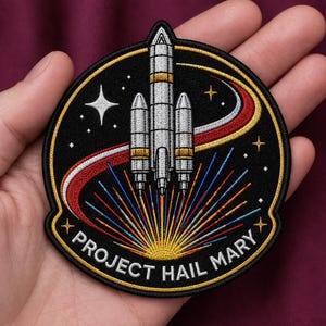 May include: A circular embroidered patch featuring a silver rocket ship launching against a black background. The design includes a red and yellow curved trail, colorful light rays at the base, white stars, and the text PROJECT HAIL MARY in white.