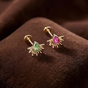 May include: Two gold stud earrings with teardrop-shaped gemstones. One earring features a green gemstone, the other a pink gemstone. Each gemstone is surrounded by a sunburst design.
