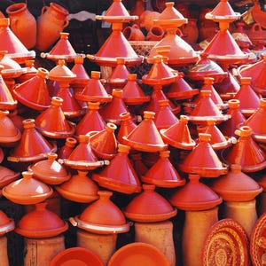 May include: A vibrant display of terracotta tagines and serving dishes. The conical lids and rounded bases of the tagines are stacked, showcasing their traditional design. The color palette is dominated by warm orange and red hues.