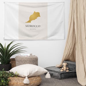 May include: A rectangular flag with a gold map of Morocco, the country's name, and "North Africa" printed in black. The flag has a white background with a vertical beige stripe in the center.