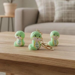Puede incluir: Tres pequeñas figuras de serpiente de color verde claro con vientre blanco y rayas horizontales de color verde oscuro. Cada serpiente tiene grandes ojos negros y una boca pequeña. Una de las figuras está unida a un llavero de metal dorado.