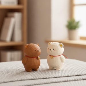 May include: Two small bear figurines, one brown and one cream-colored, each featuring a carved texture, a red string necklace with a tiny gold bell, and simple painted facial features including black eyes and a small nose on the cream bear.