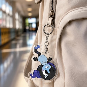 May include: A clear acrylic keychain featuring a cartoon character with a black head, blue hat, and light blue shirt, attached to a silver metal ring and chain. The character is depicted in a playful pose with one eye closed and a wide smile.