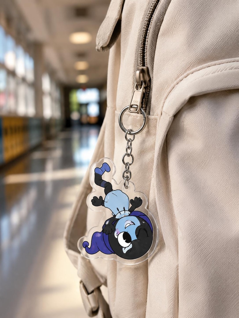 May include: A clear acrylic keychain featuring a cartoon character with a black head, blue hat, and light blue shirt, attached to a silver metal ring and chain. The character is depicted in a playful pose with one eye closed and a wide smile.