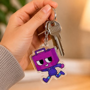 May include: A clear acrylic keychain featuring a cartoon character with a purple rectangular head, wearing a dark blue hoodie and striped leggings, attached to a silver metal split ring that also holds a standard silver house key.