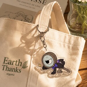 May include: A beige canvas tote bag with the text Earth Thanks organic printed in dark green. Attached to the handle is a clear acrylic keychain featuring a cartoon character with a magnifying glass head, a purple bowtie, and a black suit.