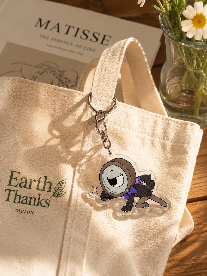 May include: A beige canvas tote bag with the text Earth Thanks organic printed in dark green. Attached to the handle is a clear acrylic keychain featuring a cartoon character with a magnifying glass head, a purple bowtie, and a black suit.