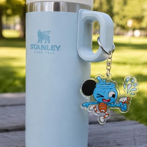 May include: A light blue Stanley brand insulated tumbler with a white lid and handle. A silver metal keychain is attached to the handle, featuring a clear acrylic charm of a cartoon character with a blue head, orange striped legs, and a spray bottle.