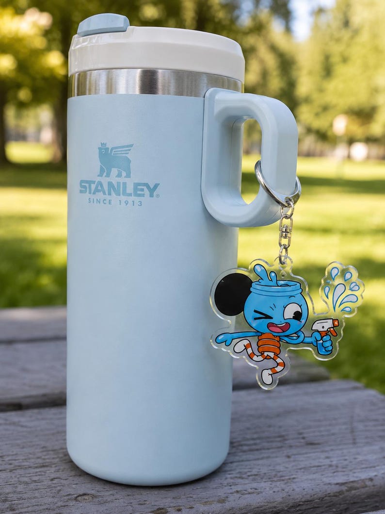 May include: A light blue Stanley brand insulated tumbler with a white lid and handle. A silver metal keychain is attached to the handle, featuring a clear acrylic charm of a cartoon character with a blue head, orange striped legs, and a spray bottle.