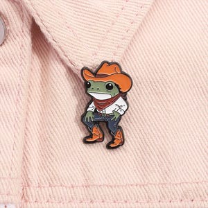 May include: A whimsical enamel pin featuring a green frog dressed as a cowboy. The frog wears a brown cowboy hat, red bandana, white shirt, blue jeans, and brown cowboy boots. The pin is set against a pink denim jacket.