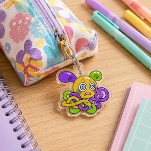 May include: A floral patterned pencil case with a zipper closure attached to a clear acrylic keychain. The keychain features a yellow cartoon character with swirling eyes and multiple purple and green tentacles, dangling from a silver metal chain.