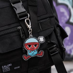 May include: A clear acrylic keychain featuring a cartoon character with a red face, a light blue beanie, and a matching hoodie, displaying an angry expression with crossed arms. The keychain is attached to a black backpack with a plastic buckle.