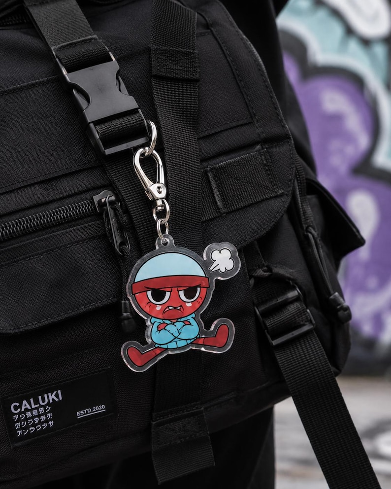 May include: A clear acrylic keychain featuring a cartoon character with a red face, a light blue beanie, and a matching hoodie, displaying an angry expression with crossed arms. The keychain is attached to a black backpack with a plastic buckle.