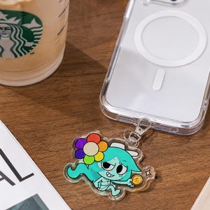 May include: A clear acrylic phone charm featuring a cartoon character with teal hair, a multi-colored flower, and a gold coin, attached to a transparent smartphone case. The charm has a glossy finish and a silver metal lobster clasp connector.