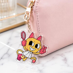 May include: A clear acrylic keychain featuring a cartoon yellow cat character with pink ears, wearing a white dress and pink gloves, holding a small pink hand mirror and making a peace sign, attached to a light pink rectangular zippered wallet.