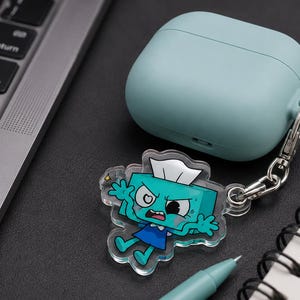May include: A light blue silicone case for wireless earbuds attached to a clear acrylic keychain charm. The charm features a cartoon character shaped like a teal tissue box with a white tissue on top, wearing a blue dress, with an angry facial expression.