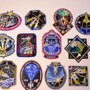 May include: A collection of eleven embroidered NASA space mission patches featuring various shapes, including circular, octagonal, and shield designs. Each patch displays colourful illustrations of space shuttles, Earth, stars, and mission text.