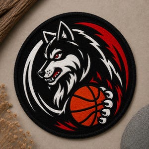 May include: A circular embroidered patch featuring a stylized wolf head in black, white, and red. The wolf has sharp teeth, red eyes, and is holding an orange basketball with black lines. The design is enclosed within a black stitched border.