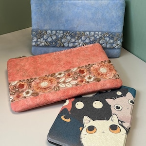 May include: Three decorative pouches in the image. The top pouch is light blue with floral embroidery. The middle pouch is coral with floral embroidery. The bottom pouch is a tablet case with a cat illustration.