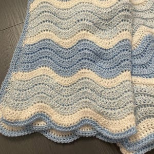 Puede incluir: Una manta de crochet hecha a mano en tonos azul claro y crema. La manta presenta un patrón ondulado, con cada ola de un color diferente. Los bordes están festoneados, agregando un toque decorativo. Esta suave manta es perfecta para un bebé.