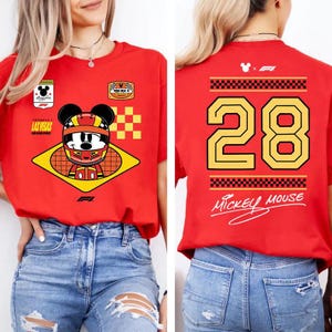May include: A bright red t-shirt featuring a graphic of Mickey Mouse wearing a racing helmet on the front, with the text Las Vegas Grand Prix. The back displays the number 28 in large yellow font, checkered patterns, and the text Mickey Mouse.