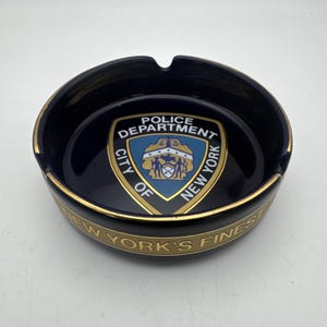 Puede incluir: Una taza de cerámica blanca con asa negra y el texto "But First, Coffee" en negrita negra. La taza se destaca sobre un fondo borroso, resaltando su diseño limpio.