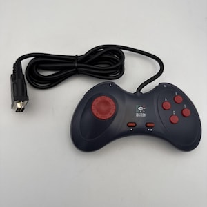 May include: A black Logitech gamepad with a red directional pad and four red action buttons. The controller has a black cable and a connector on the end. The Logitech logo is visible on the controller.