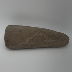 May include: A brown, oblong stone tool with a rounded end and a slightly tapered shape. The surface has a rough texture and a diagonal line carved into it. The tool appears to be an ancient artifact, possibly used for grinding or other tasks.