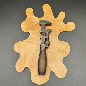 May include: An antique adjustable wrench with a dark wooden handle and a metal head. The wrench is set against a tan leather backdrop, showcasing its vintage design and craftsmanship. The tool is likely used for tightening or loosening bolts.