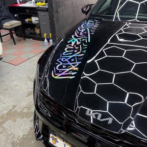 Puede incluir: Un coche negro con un diseño holográfico en el capó. El coche tiene una matrícula de Nueva York con las letras TJK. El logotipo de Kia es visible en la parte delantera.